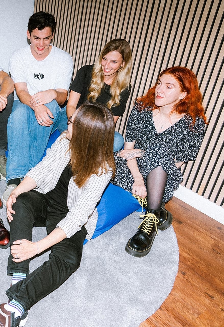 Four young adults are sitting in a circle on blue beanbags in front of a wooden slatted wall, laughing.